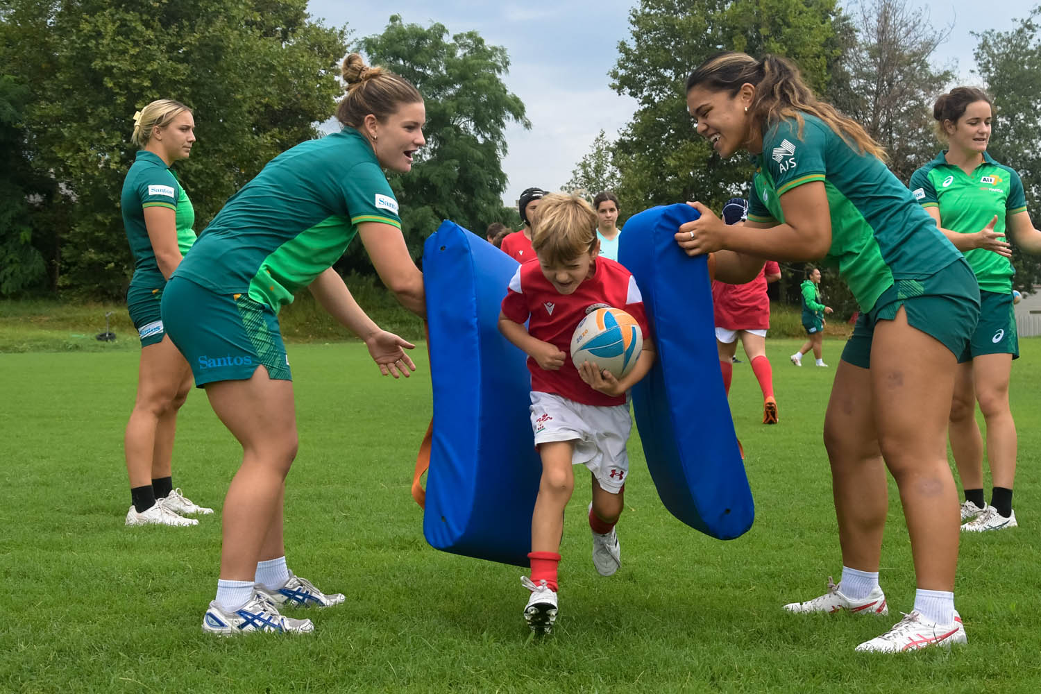 A young boy holding a rugby ball runs between two Australian rugby 7s players hold up tackle pads.