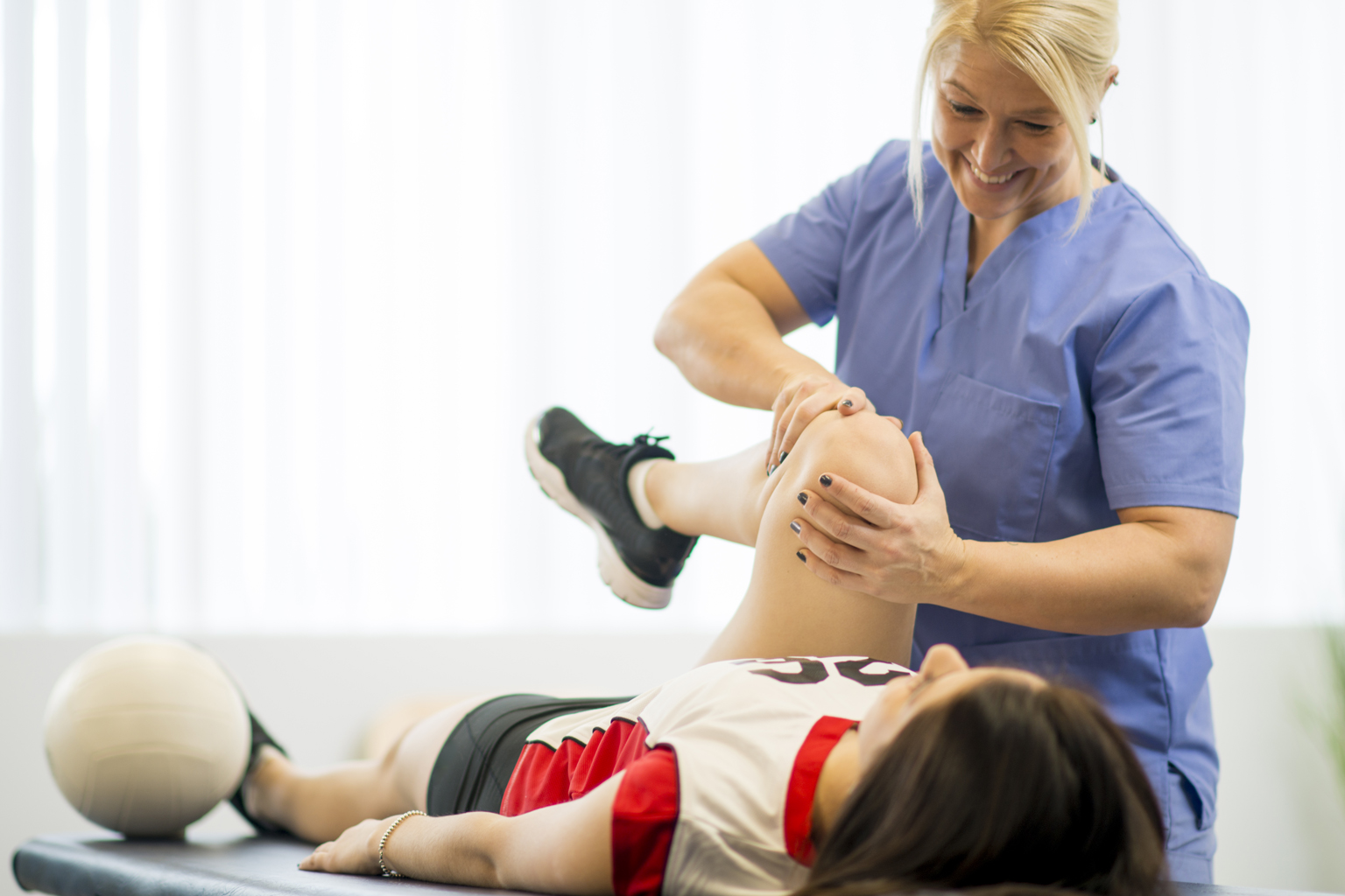 A sports therapist helps holds an athlete's knee while she lays on a treatment bed, helping her stretch her hamstring.