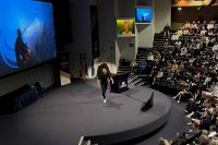 A man stands on stage, speaking, with a screen showing an image of a surfer