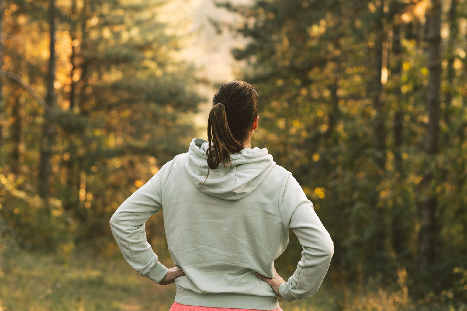A woman in a hoodie looks out towards forested area with hands on hips. 