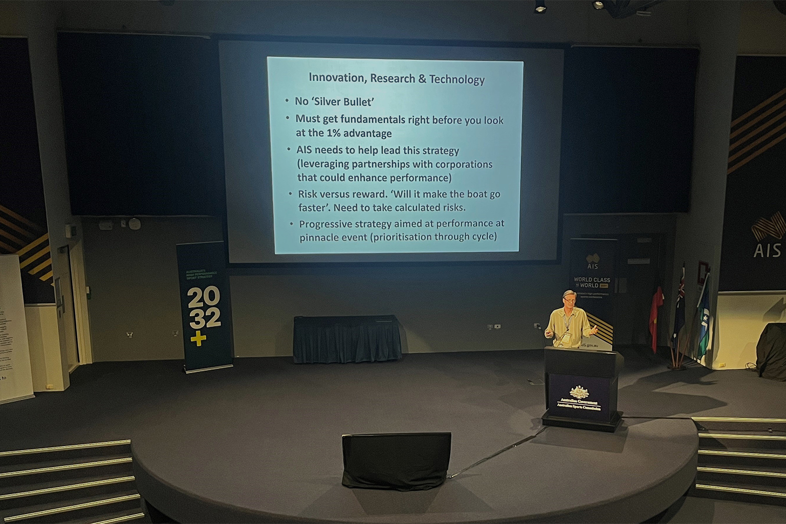 A man on stage with a large screen of text under the heading 'Innovation, Research and Technology' in the background