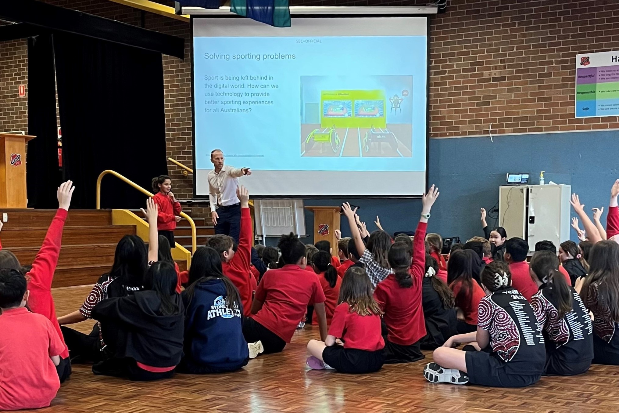 Students sit on gym floor with hands up, looking at screen.