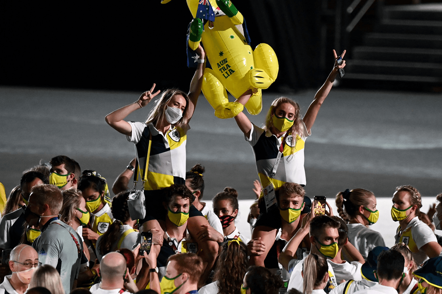 Australian athletes at the Tokyo 2020 Olympic Games closing ceremony, including two young women sitting on male athlete's shoulders, holding an inflatable boxing Kangaroo and making peace signs.