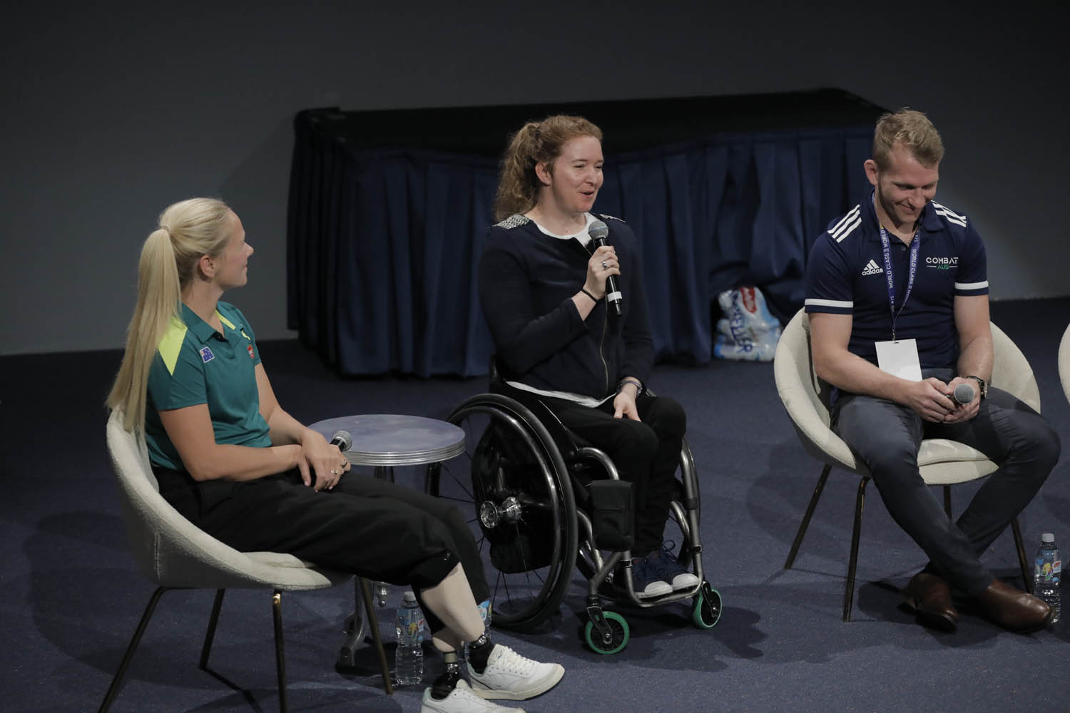 A woman with prosthetic legs, a woman in a wheelchair and a man seated on stage