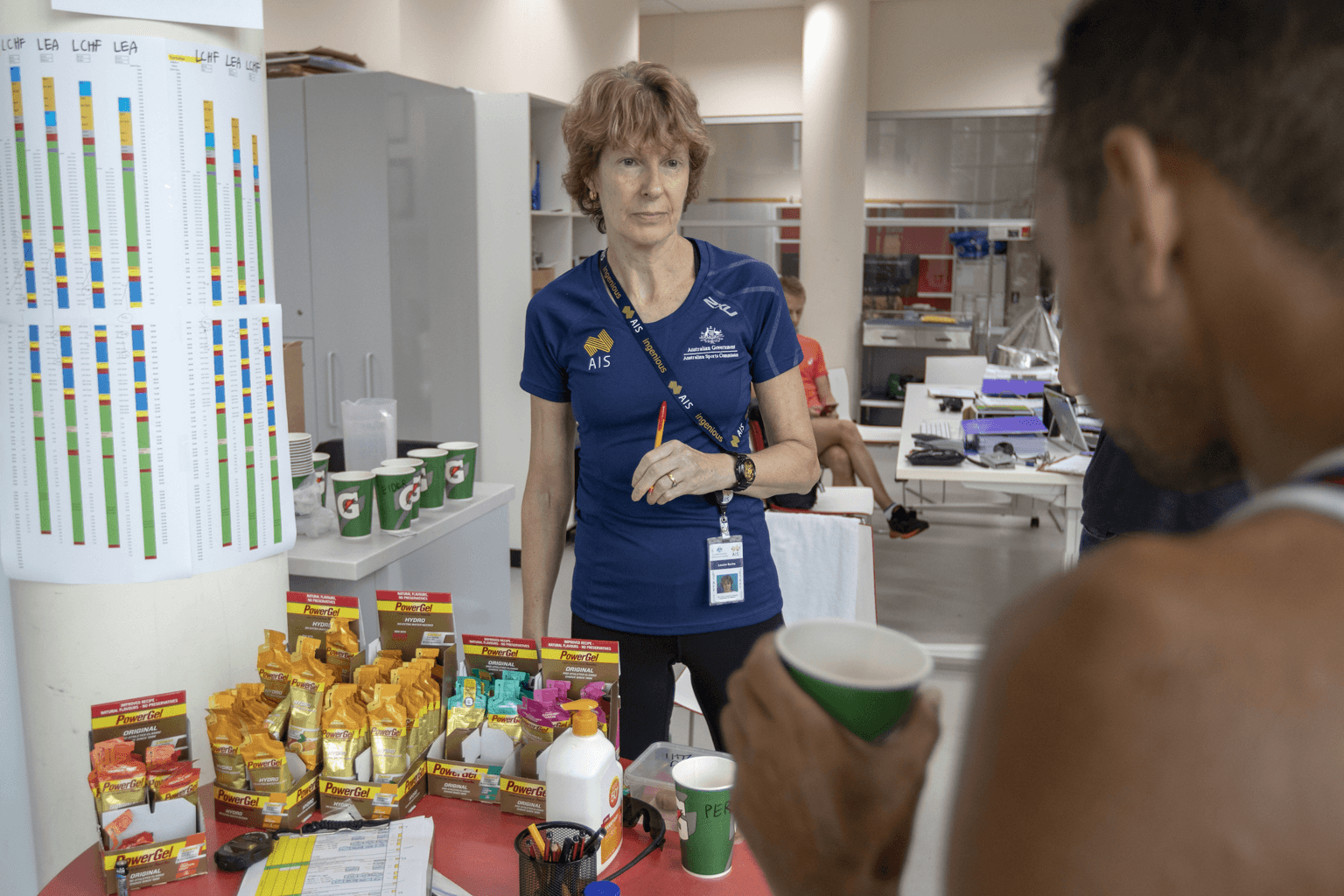 Professor Louise Burke stands behind a table with sport supplements and papers, while and athlete drinks from a cup.