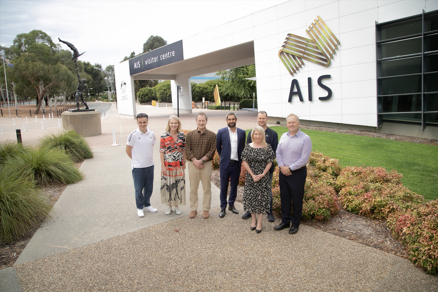 A group of people standing outside the AIS Visitor Centre, with a sculpture and gardens in the background.