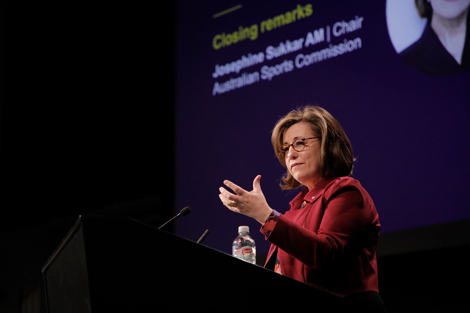 A woman gesturing behind a lectern, speaking into a microphone