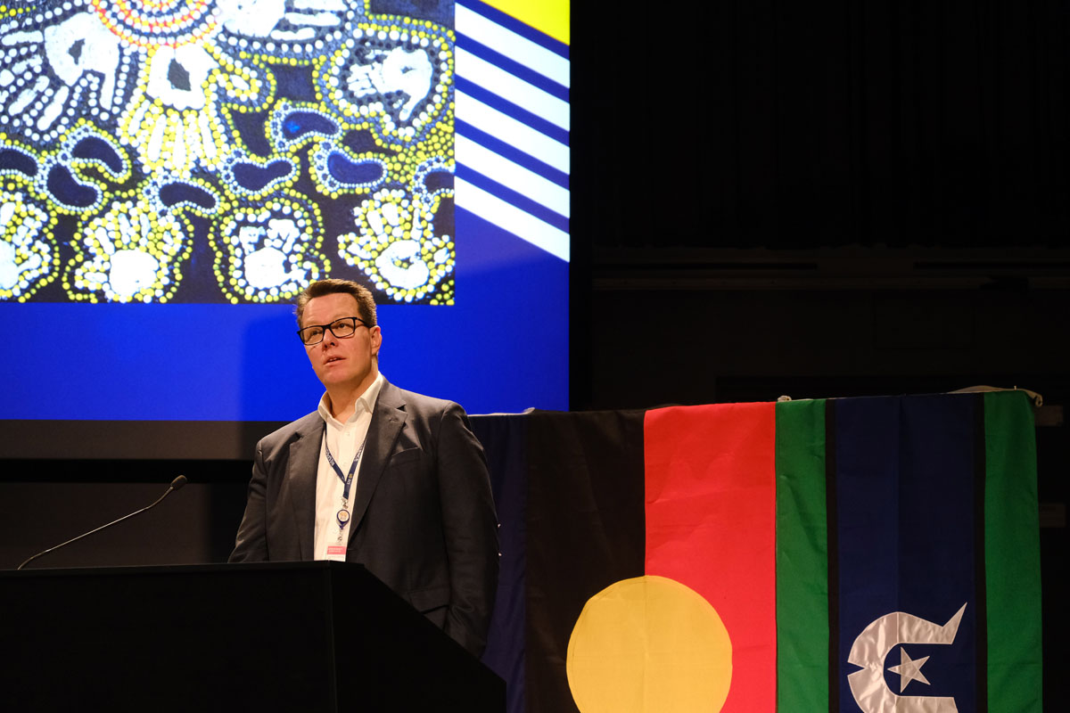 Kieren Perkins stands below an Aboriginal artwork projected onto the wall behind him