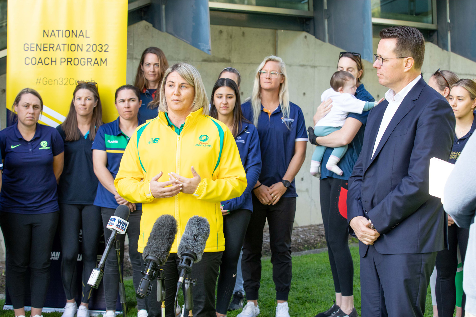 Woman in yellow Diamonds netball jacket speaks into microphones with a group of women coaches in the background.
