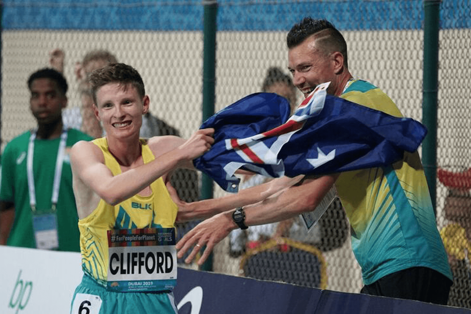 Jaryd Clifford wears a racing bib and waves an Australian flag with coach Philo Saunders