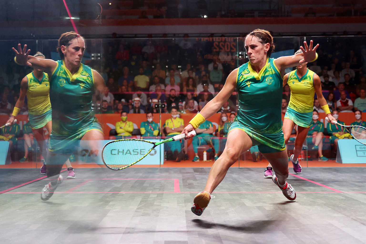 A squash player reaches for the ball, with her reflection clearly visible in the court's perspex wall. 