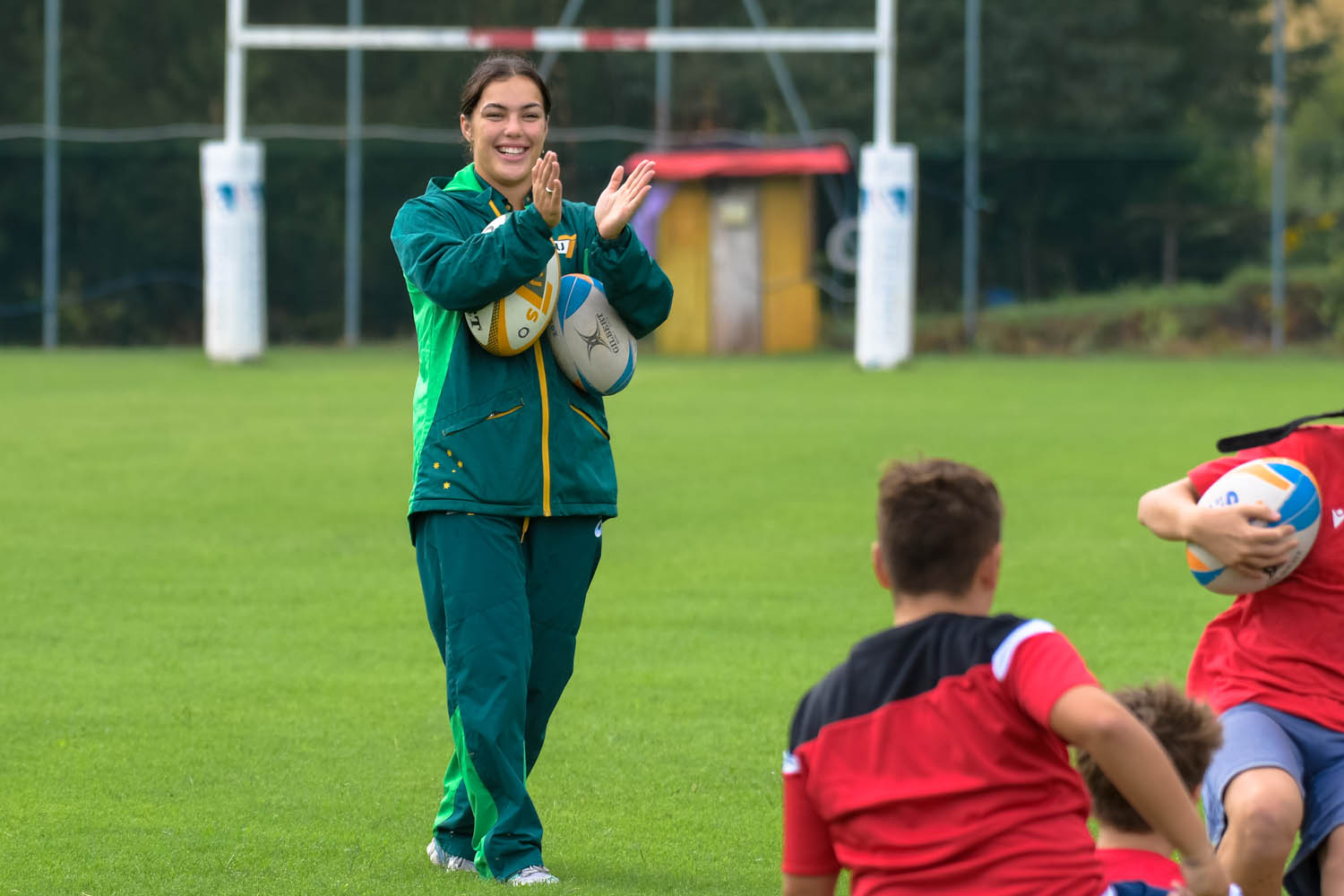 A woman claps for junior rugbyy players while holding two rugby balls to her body.